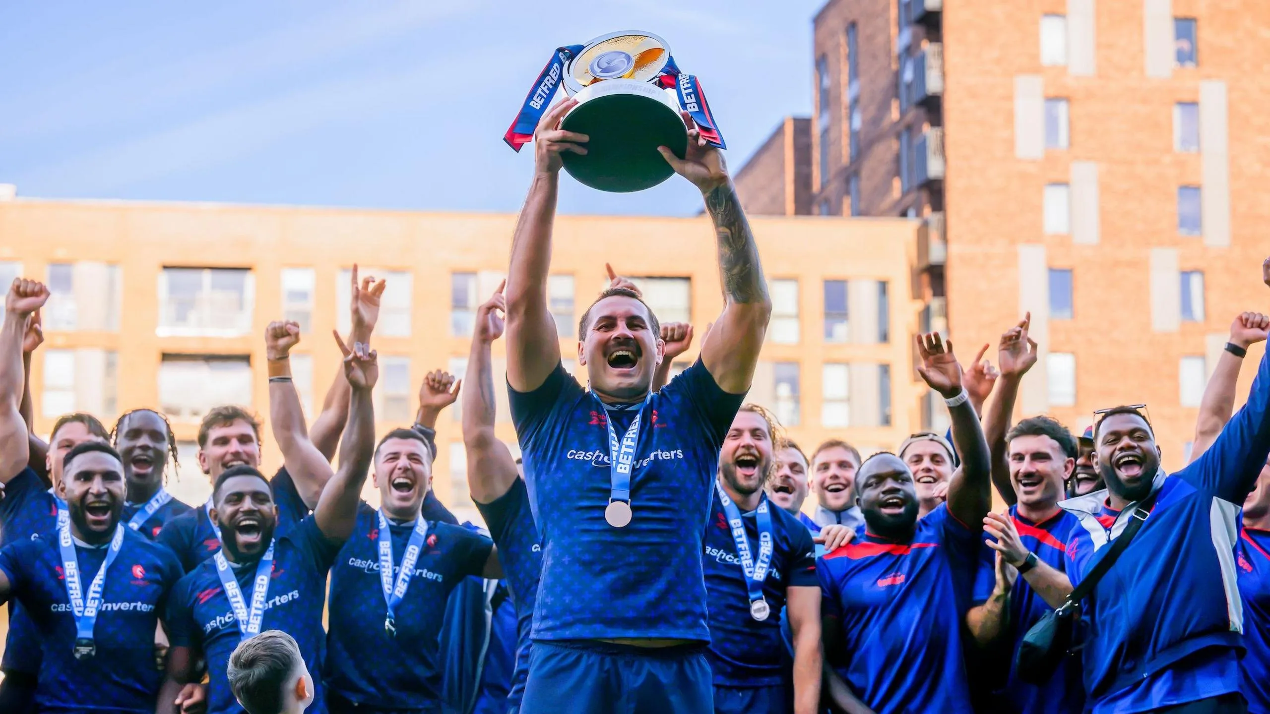 The captain of London Broncos lifts the Championship trophy as his team-mates lift their arms in celebration behind him