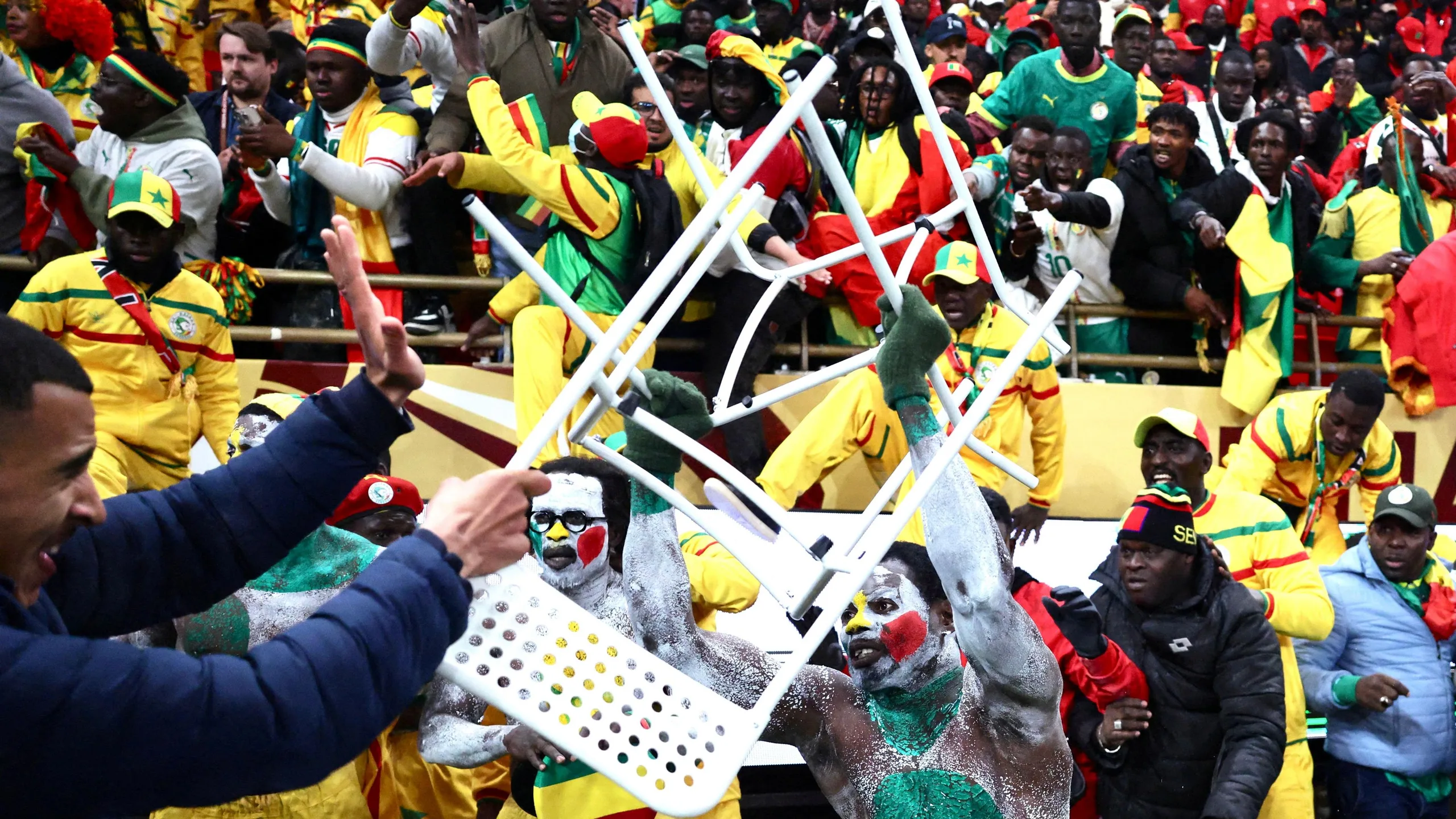 Senegal supporters clash with security personnel as they attempt to storm the field of play after a penalty decision against their team during the 2025 Africa Cup of Nations final in Rabat. In the central foreground is a man covered in white body paint with green, yellow and red dots launching a white chair at a man in a blue puffer jacket who is grimacing, holding up his left hand in an effort to protect himself while also wielding a similar chair with his right hand. Behind the two central characters are several other Senegal supporters, some wearing all-yellow tracksuits and baseball caps, who have come out of the stand and are coming over hoardings to reach the ground. In the background is the packed stand full of Senegal supporters, many of whom are gesticulating in an animated way