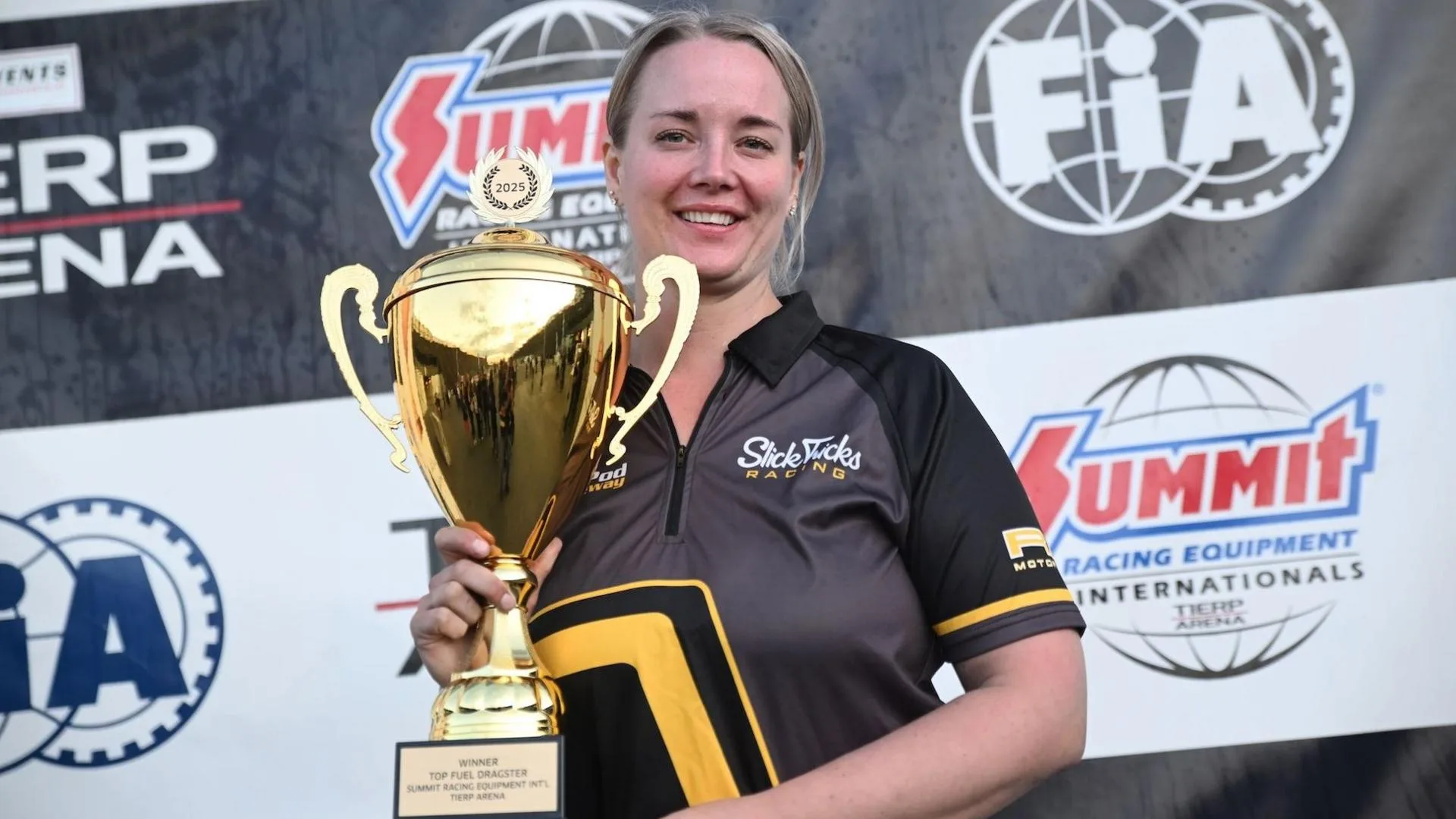 Susanne Callin holding a trophy and smiling after winning the European Top Fuel Championship in 2025 in Tierp, Sweden. Her hair is tied back and she is wearing a black shirt with yellow markings on it. She is standing in front of a black and white banner with racing logos on it.
