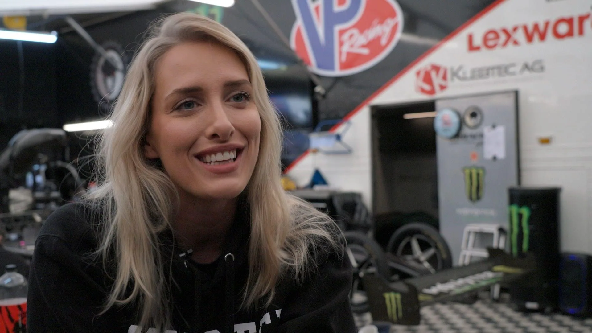 Jndia Erbacher speaking at Santa Pod drag strip near Northampton, UK. She is wearing black, has blonde hair and is smiling. Car parts and and walls with motoring logos on them can be seen behind her.