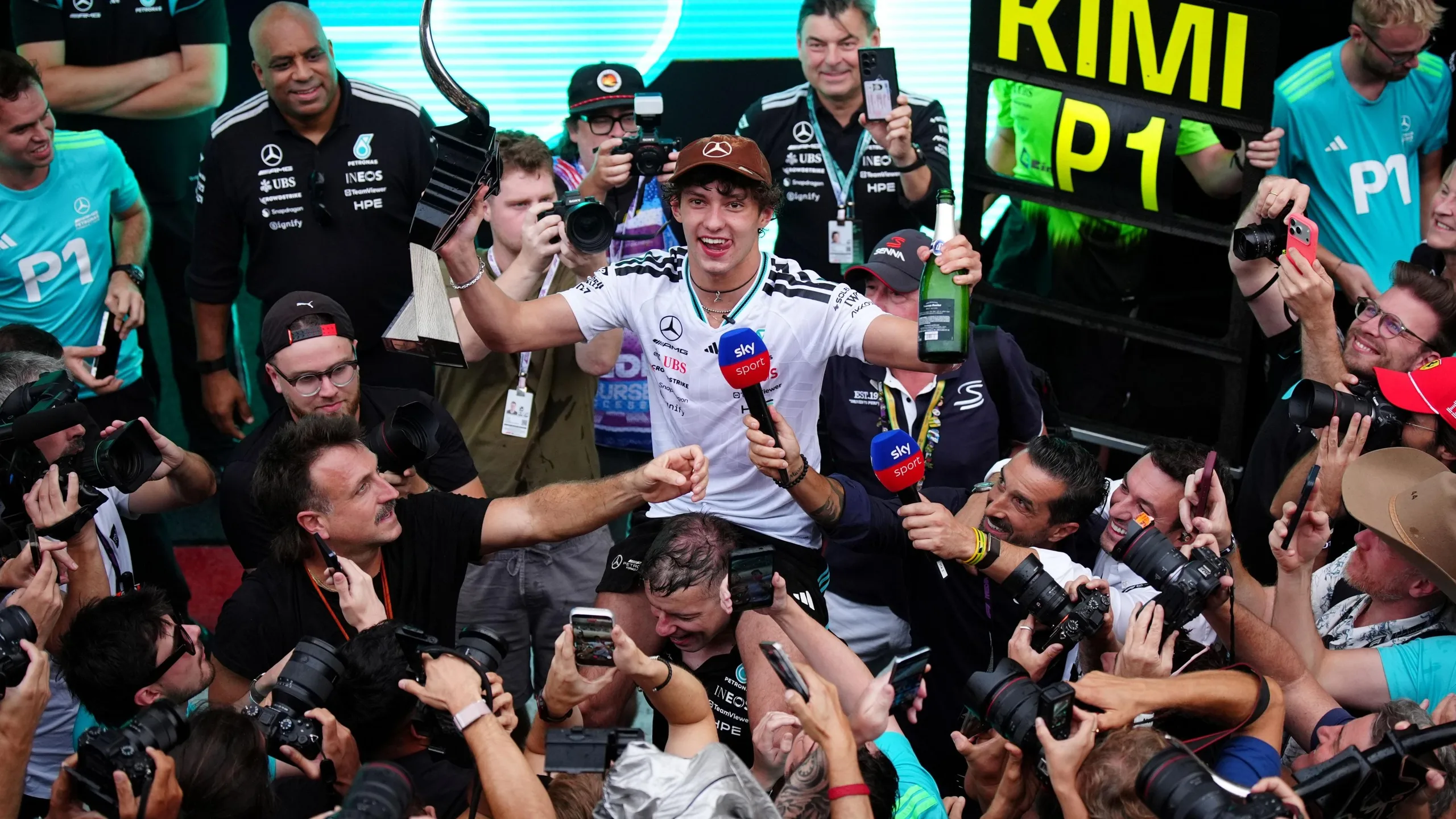 Kimi Antonelli celebrating with the Mercedes team after winning the US Grand Prix. He is surrounded by people taking photographs or holding microphones