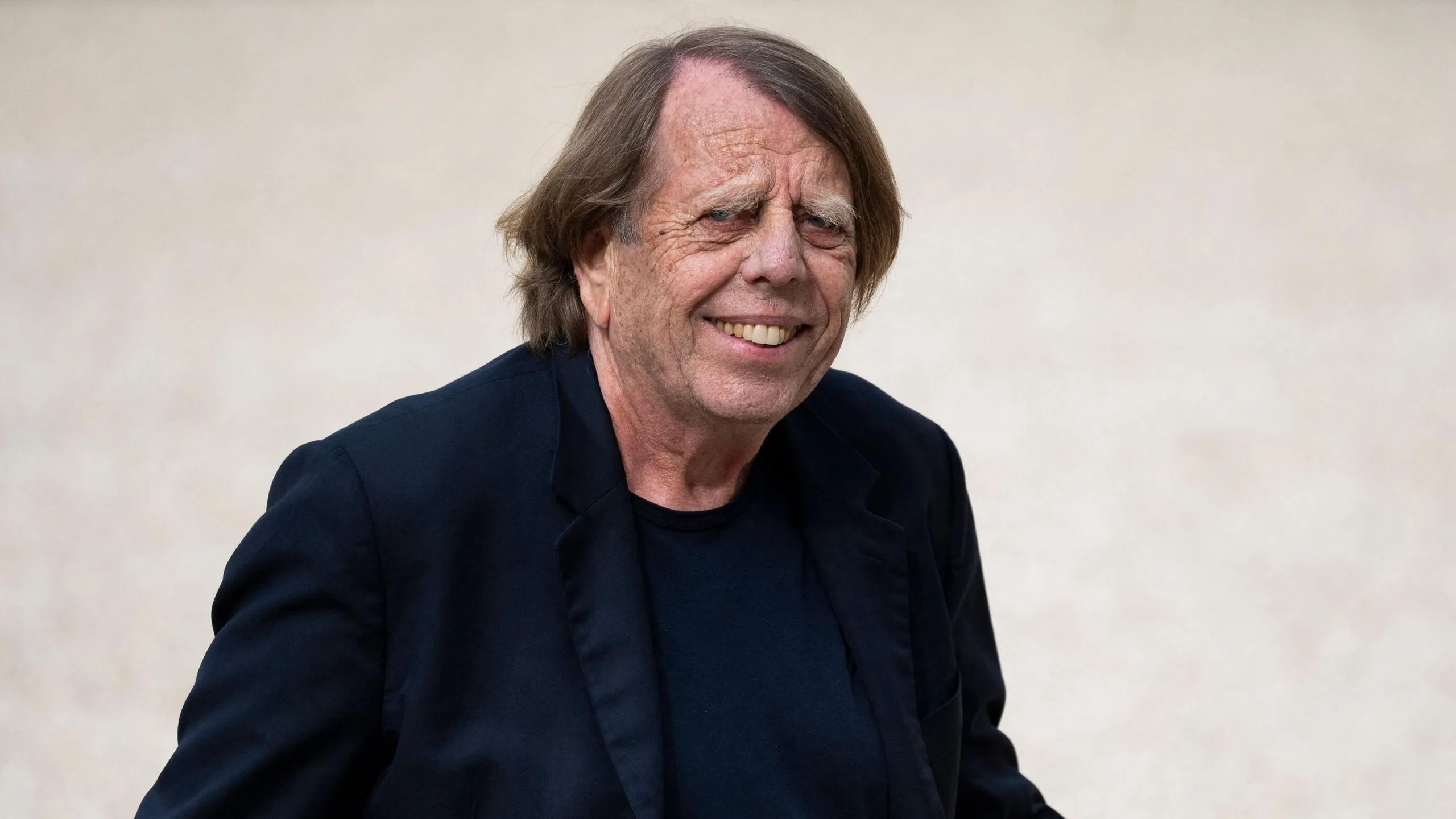 Claude Le Roy, visible from chest up wearing a dark blue jacket over a dark blue jumper, glances to his right and smiles while walking