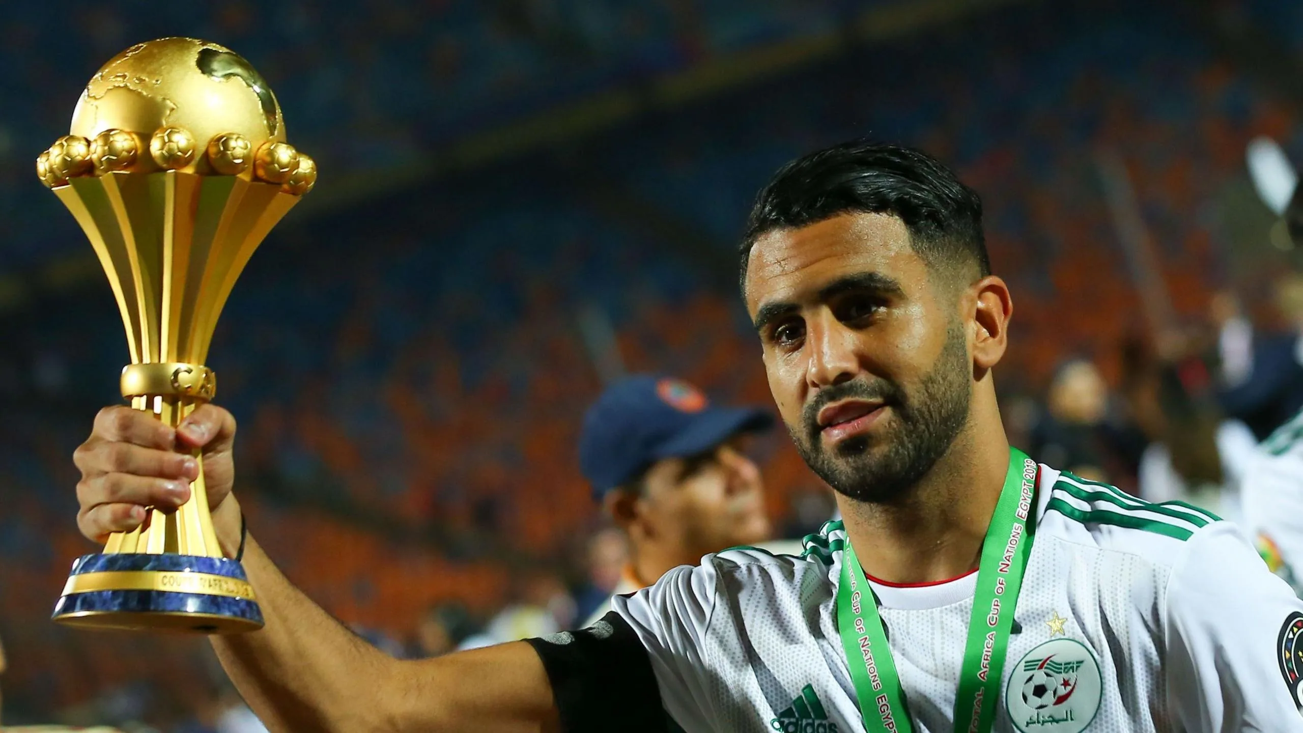 Riyad Mahrez, visible from shoulders up wearing a white Algeria shirt with a green ribbon around his neck, holds up the golden Africa Cup of Nations trophy in his right hand. A predominantly empty football stand can be seen out of focus in the background while other people mill around, making it clear Mahrez is on a pitch post-match