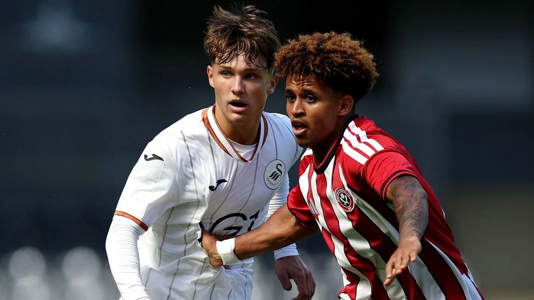 Siem Eyob-Abraha, visible from waist up wearing a Sheffield United shirt with red and white vertical stripes, holds off Swansea City's Kai Rhodes, who is wearing a white shirt with thin bronze vertical pinstripes. Eyob-Abraha is using his right hand to grab a portion of Rhodes' shirt around the stomach while he also stretches his left arm out in front of himself