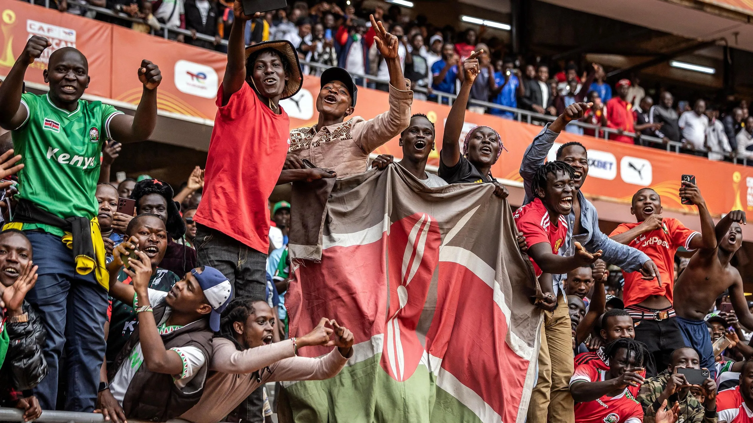 Six men stand on a railing behind a Kenyan flag as a sea of people take pictures, cheer and shout encouragement in a packed stand at the Kasarani Stadium during a match between Kenya and Morocco at the 2024 African Nations Championship