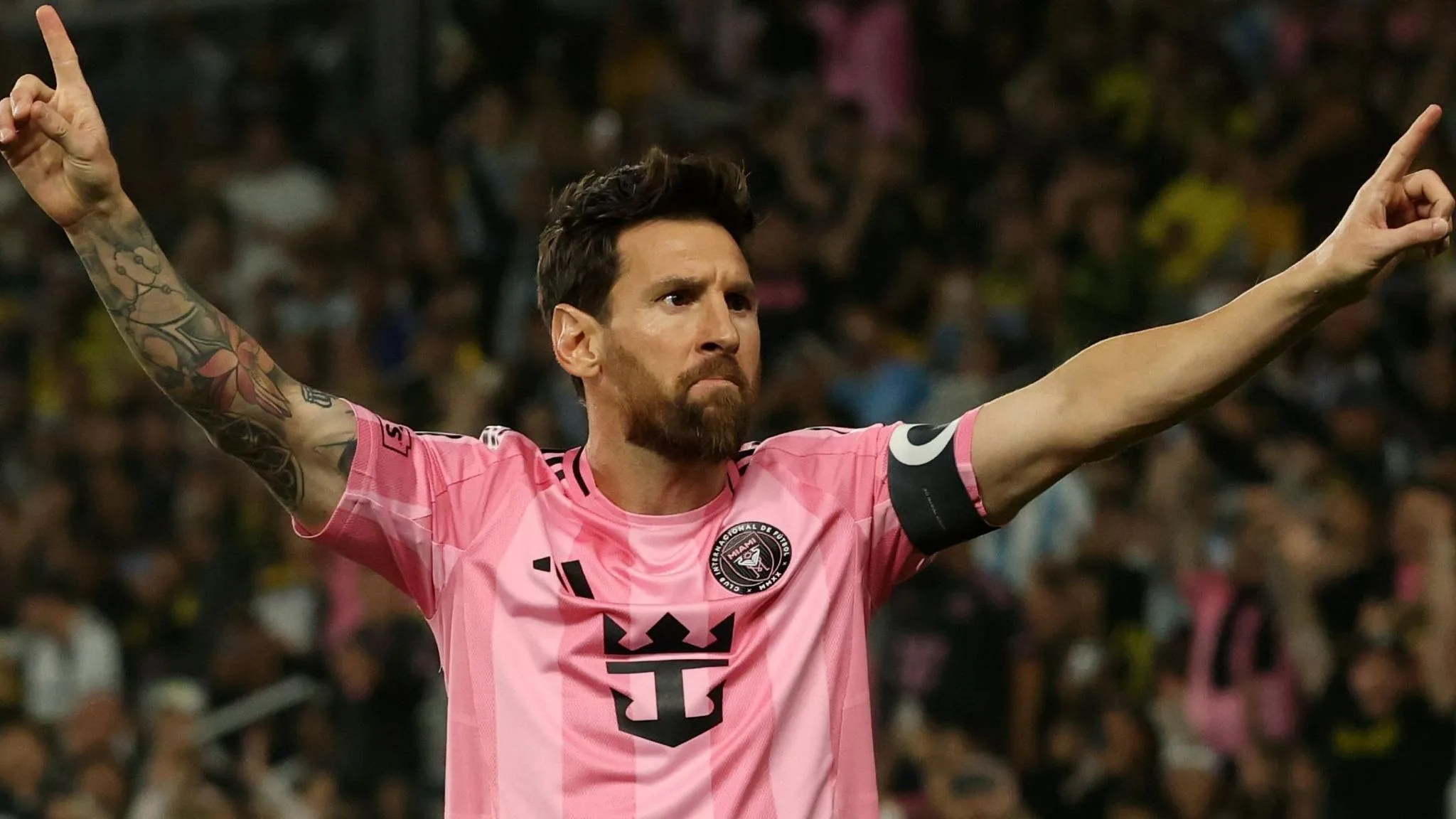 Lionel Messi celebrates while playing for Inter Miami in the MLS, wearing their pink kit and stretching out his arms upwards with index fingers pointing