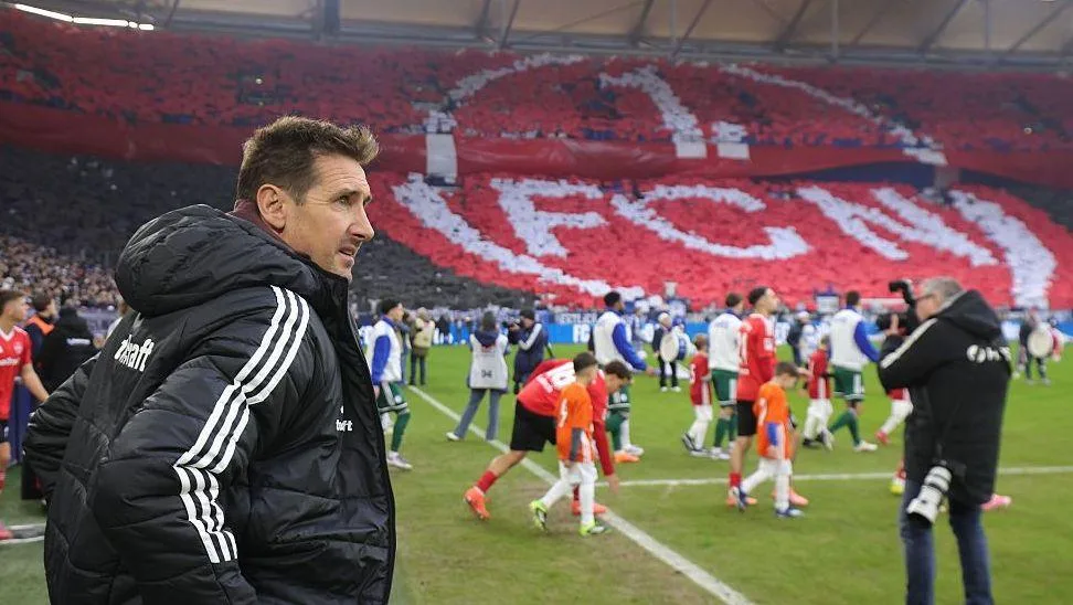 Miroslav Klose stands on the touchline in a black sports coat with three white stripes down its right sleeve