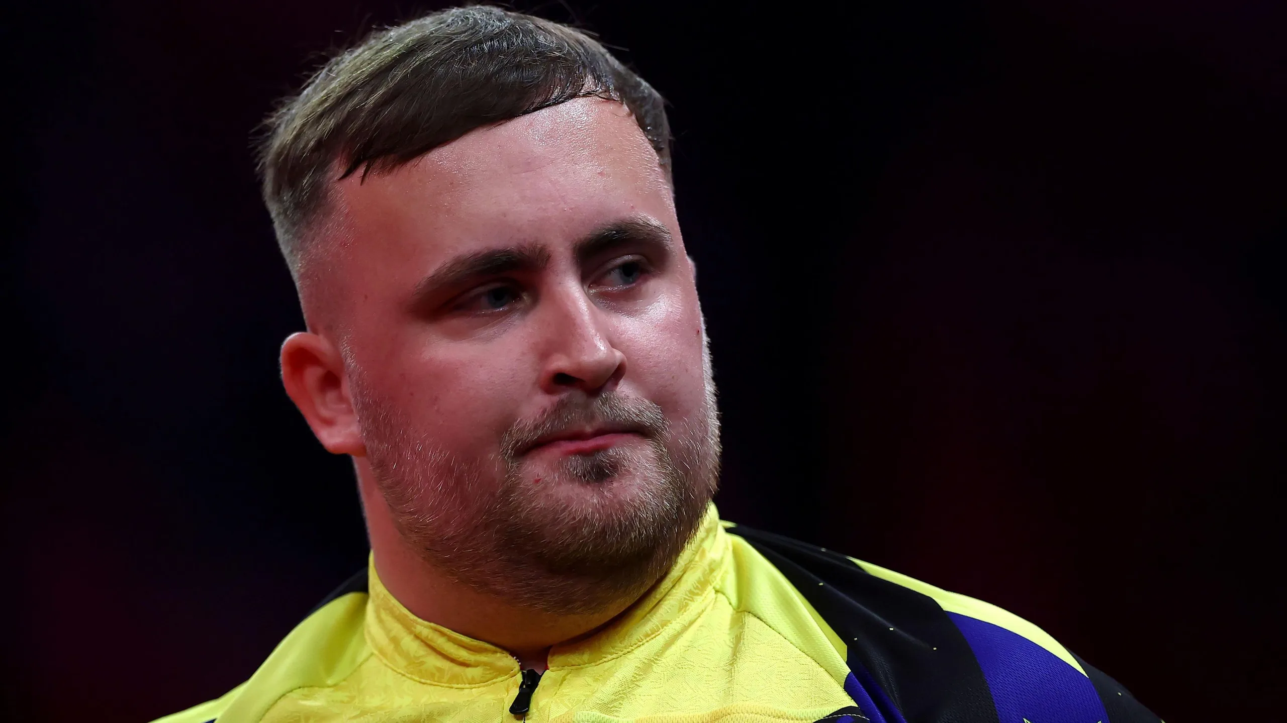 Luke Littler looking to his left in a yellow and blue darts top