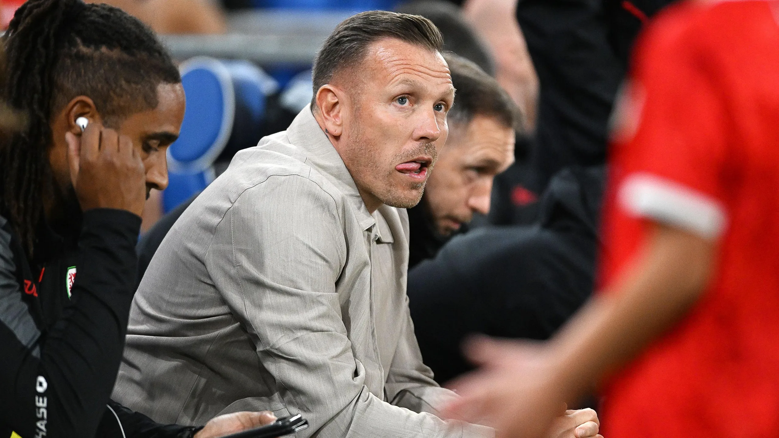 Craig Bellamy in a grey lesiure suit sits in the Wales dug out and looks towards a Wales player in the foreground