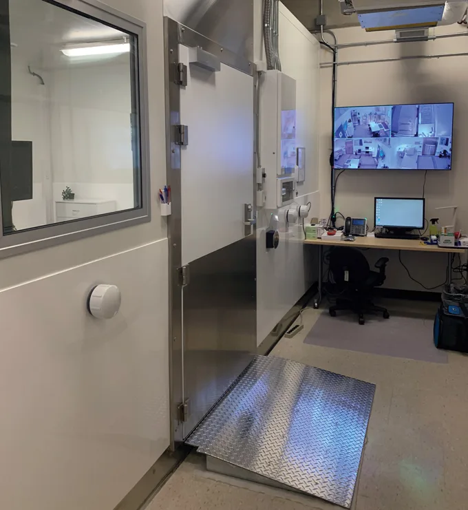 Inside the lab at the Human and Environmental Physiology Research Unit at the University of Ottawa. The left wall has a door and window looking into the room next door. Along the back wall sits a desk with a computer monitor and, above it, a larger monitor showing live images from four cameras.