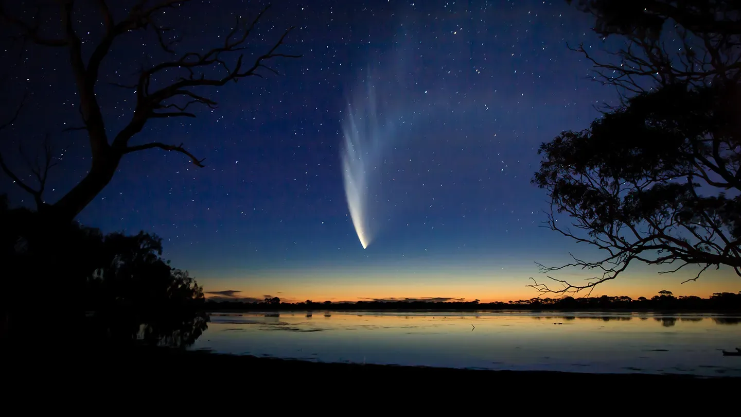 A comet's long tail arcs over a sunset over a lake, framed by trees.