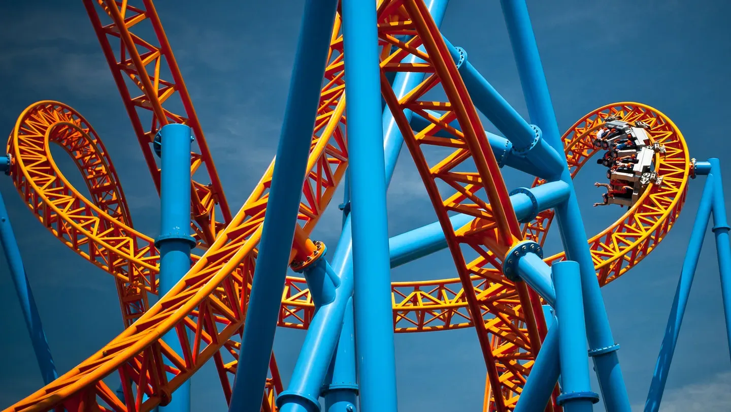 An image of a orange and teal twisting metal rollercoaster with a grey rollercoaster car full of riders