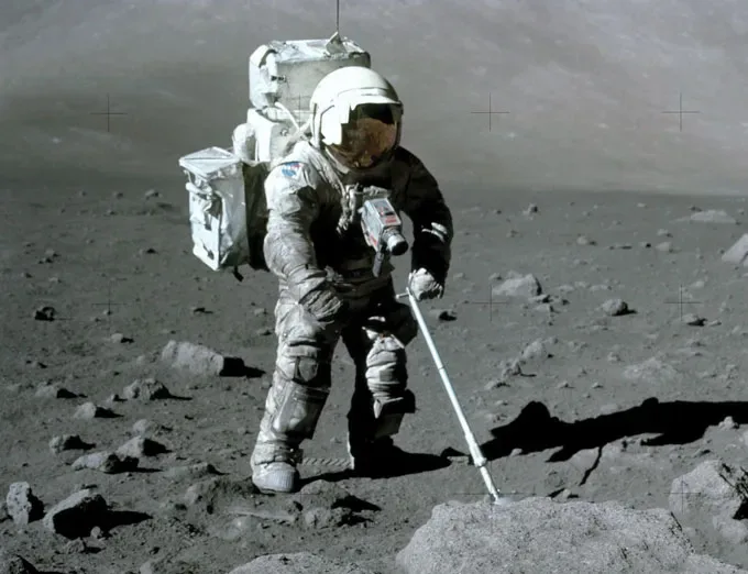 An astronaut in a bulky spacesuit stands on the rocky surface of the Moon, using a handheld tool to collect a sample. The gray, dusty landscape stretches into the distance, with scattered rocks and the stark horizon.