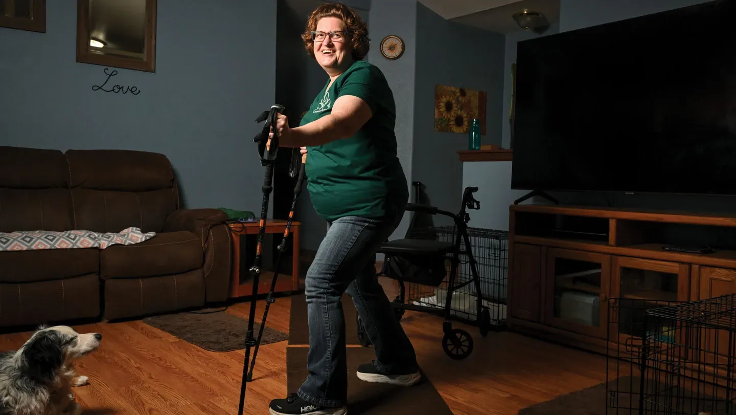 A smiling person with short, curly brown hair and glasses stands outdoors, holding a trekking pole in each hand. They wear a black top and blue jeans, with trees and buildings blurred in the backgroun