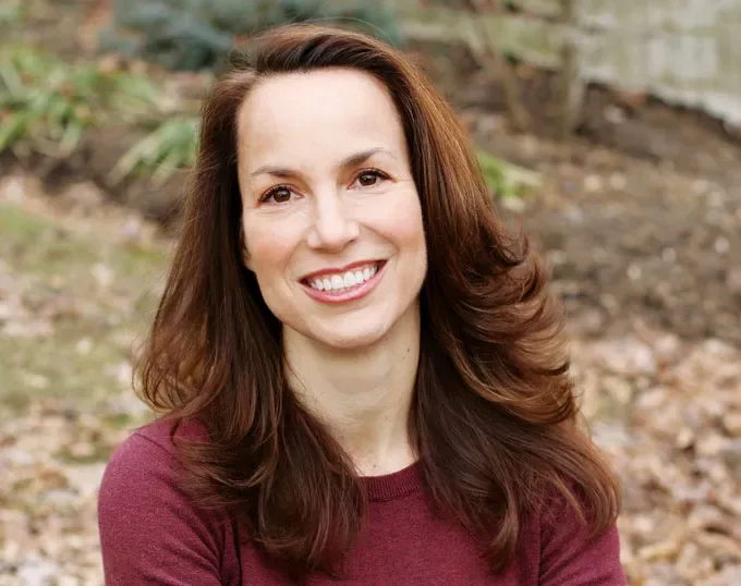 A smiling woman with long, wavy brown hair wears a burgundy sweater and looks toward the camera, with a softly blurred wooded background.