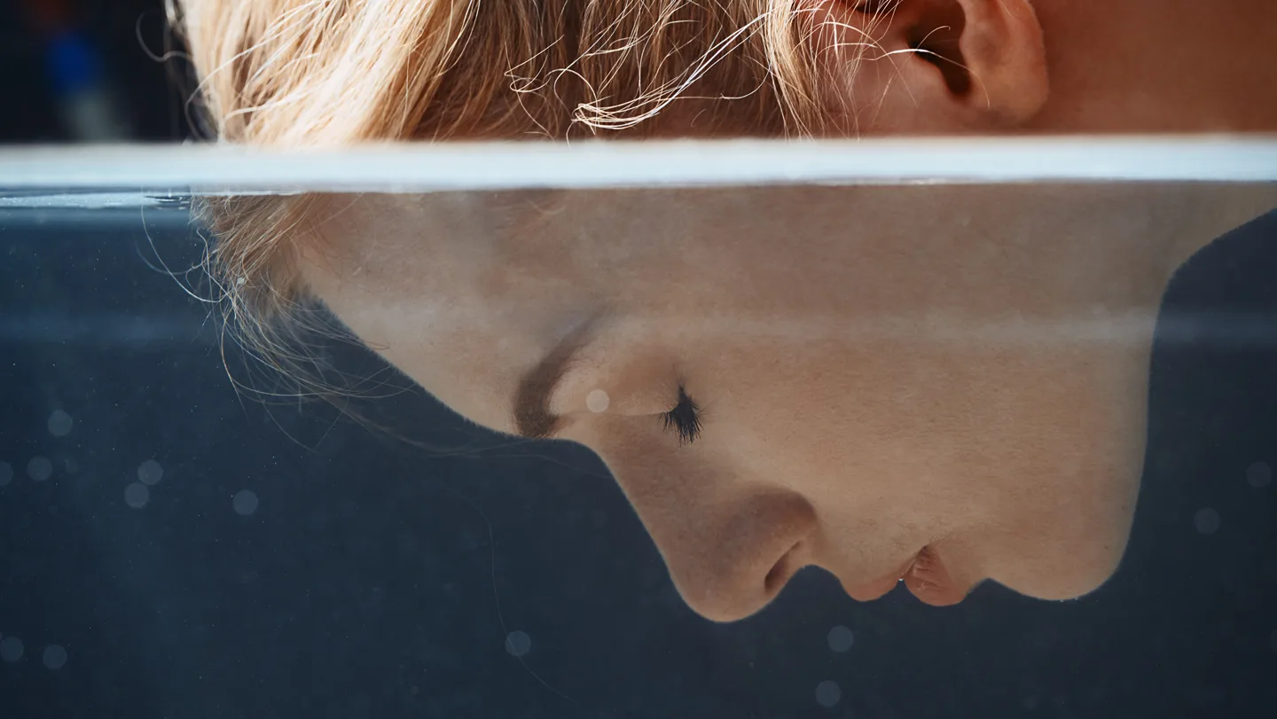 Close-up of a woman with her face partly submerged in water, her eyes closed beneath the surface.