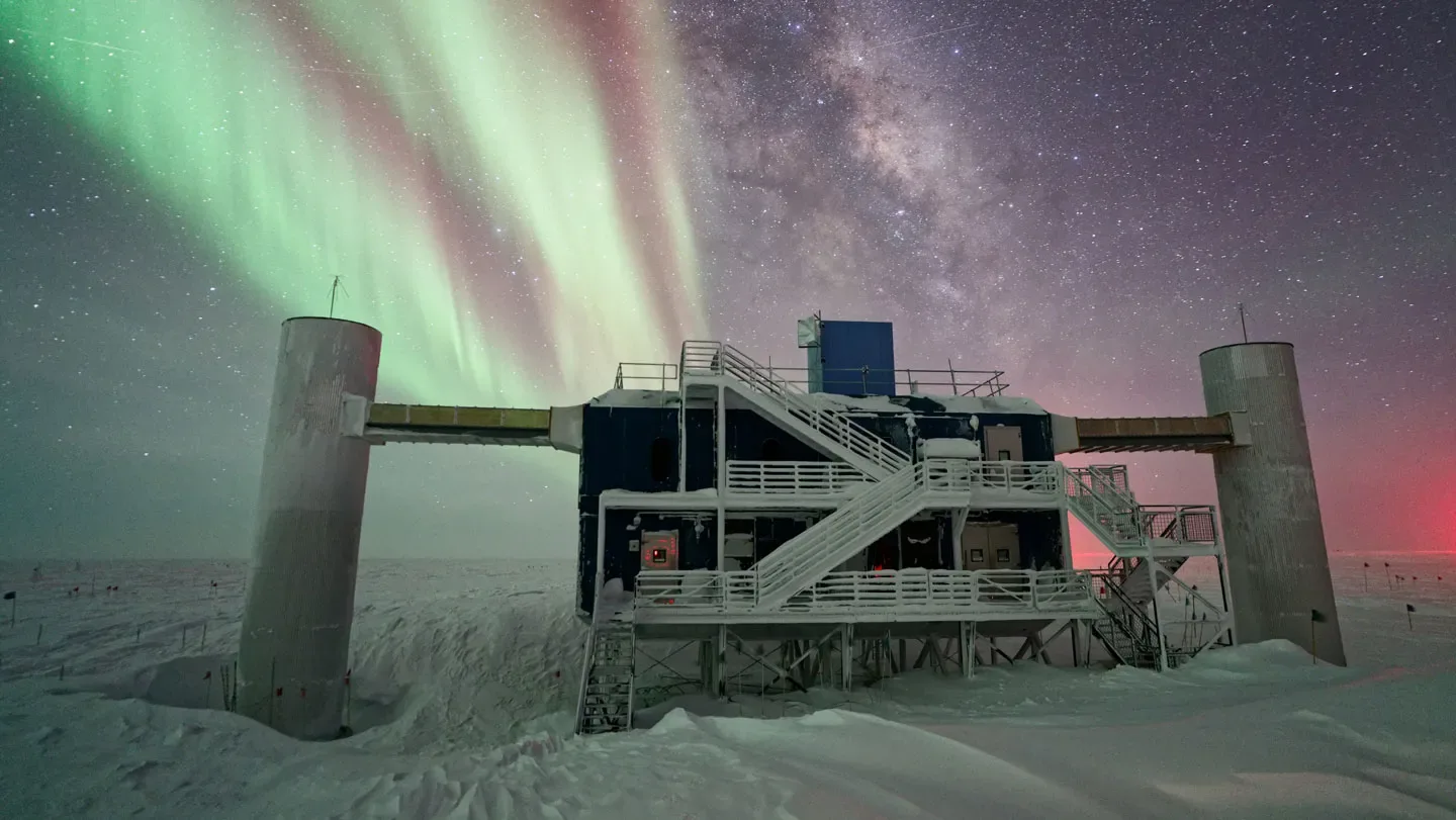 Beneath a brilliant aurora in a night sky, a building stands amid a field of snow and ice.